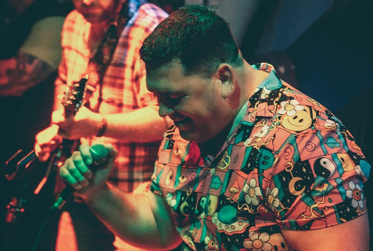 Man singing on stage wearing a colourful Bad Shirt Club party shirt with retro and psychedelic prints during a live music performance.