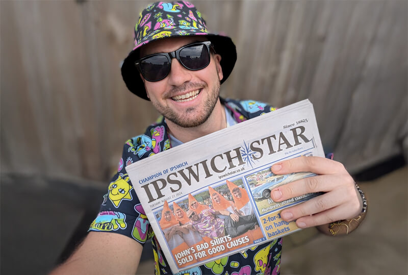A smiling man in colorful, cartoon-patterned clothes and a bucket hat holds up an Ipswich Star newspaper. He is wearing sunglasses and standing outdoors in front of a wooden fence.