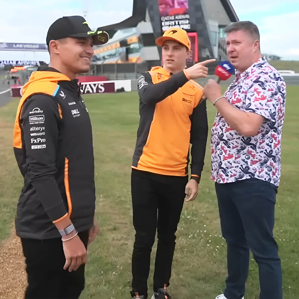 Three men stand on a racetrack; two wear orange and black racing team jackets, one holding a microphone to the other’s face, while a third man in a flowered shirt holds a microphone. The scene is casual and outdoors.