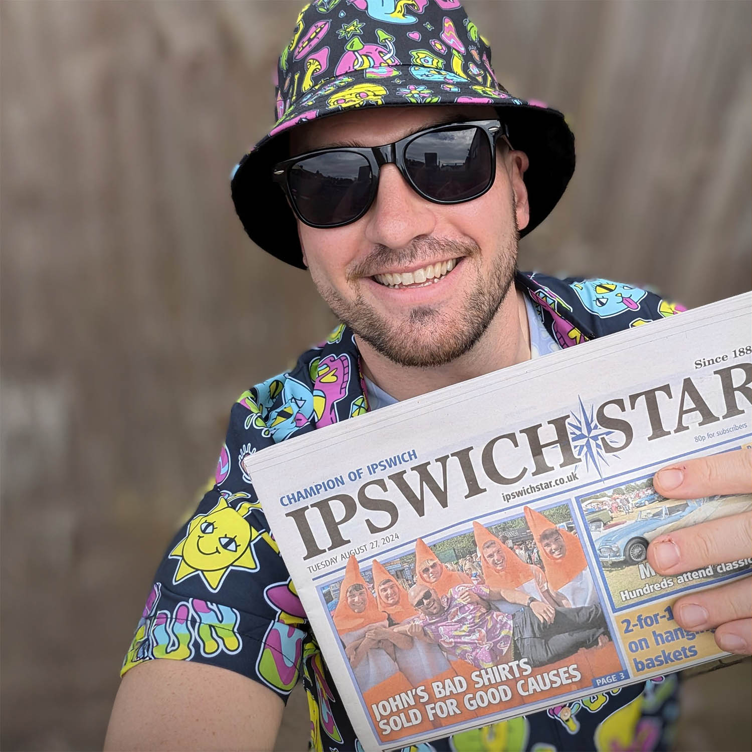 A smiling man in sunglasses and a colorful, cartoon-themed shirt and bucket hat holds up a copy of the Ipswich Star newspaper featuring a front-page story titled Johns Bad Shirts Sold for Good Causes.
