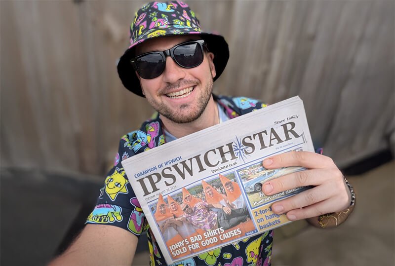 Bad Shirt Club Founder, John Parnell, holding a newspaper stating 'Johns Bad Shirts Sold For Good Causes'