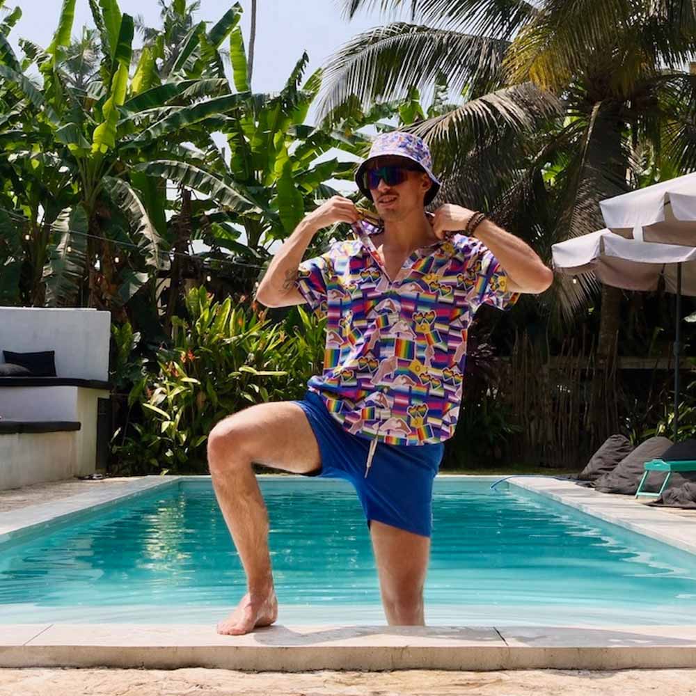 A man, dressed in a colorful shirt, blue shorts, and the LGBTQ Pride Bucket Hat, poses confidently by a pool amid lush tropical plants and palm trees on a sunny day.
