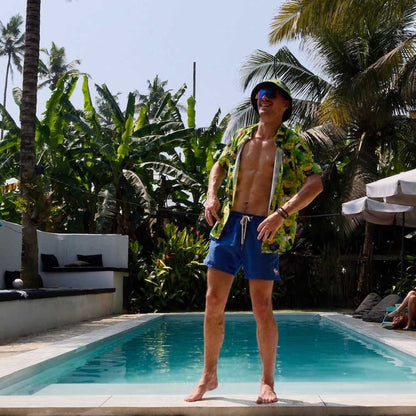 A man in a colorful shirt, blue swim shorts, and a St Patrick’s Day Bucket Hat stands confidently at the edge of a pool surrounded by palm trees and tropical plants on a sunny day.