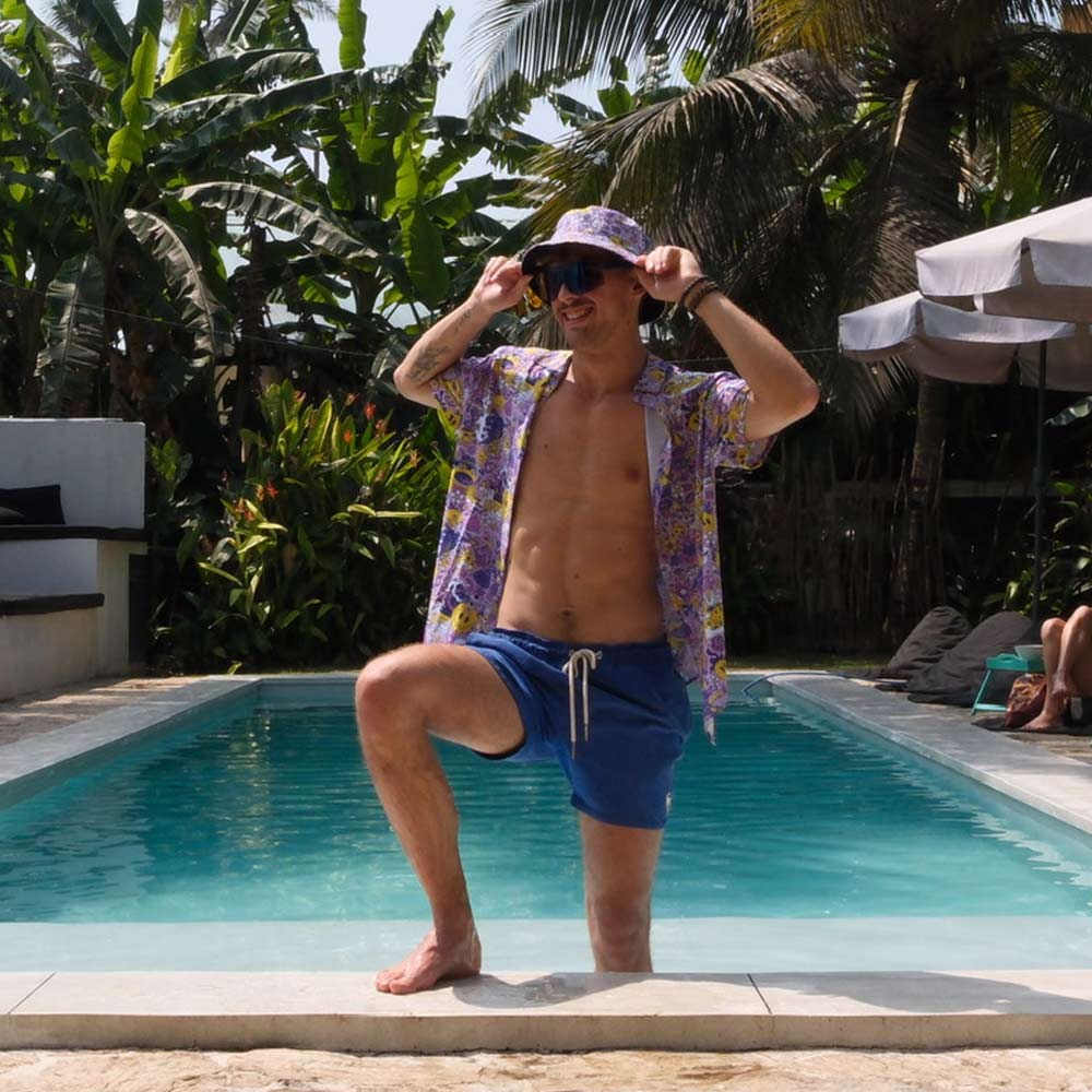 A man wearing blue swim trunks, a colorful shirt, and the Magical Psychedelic Bucket Hat kneels by the pool, smiling and adjusting his hat. Tropical plants surround him, creating an ideal festival fashion vibe.