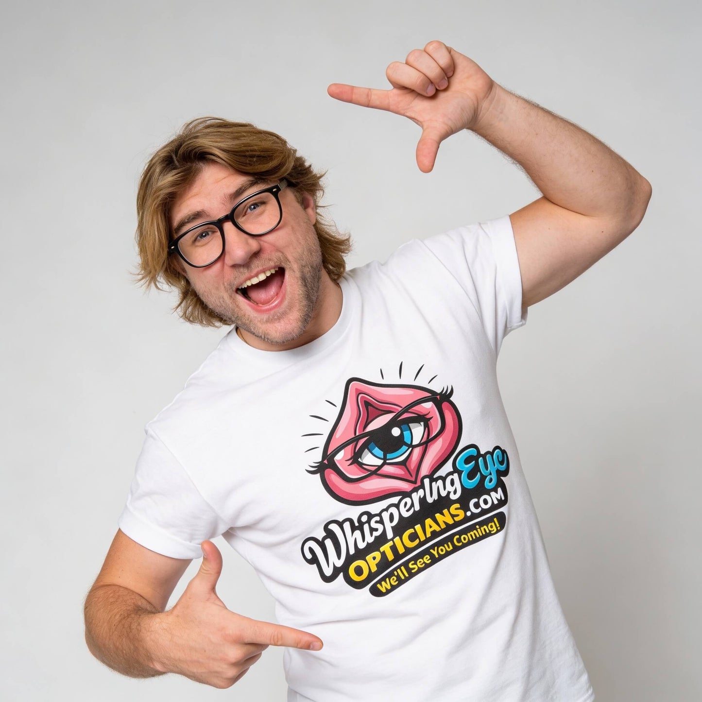 A smiling man with glasses and medium-length hair points at his Whispering Eye Opticians Logo Tee (We'll See You Coming!), which features a cartoon eye inside lips and the brand’s name on the front.