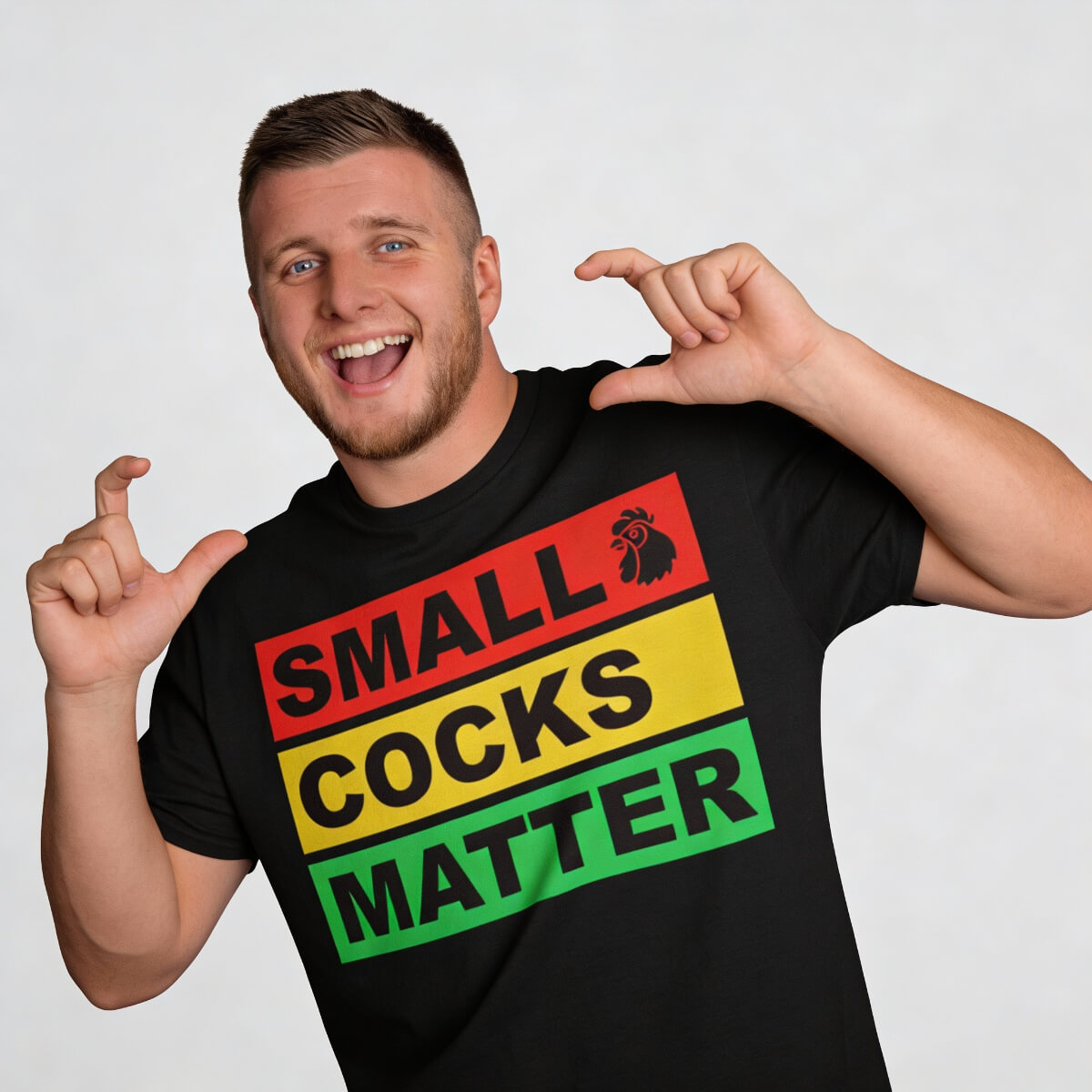 A smiling man wears the Small Cocks Matter Tee, featuring bold red, yellow, and green blocks. He gestures to show something small—an ideal funny stag party t-shirt for fans of cheeky stag do humour.
