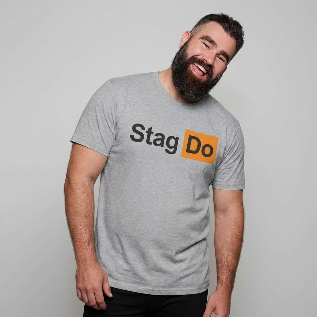 A bearded man in a gray Stag Do PornHub Tee smiles and tilts his head to the side against a plain light background, the humorous shirt adding a playful touch to his relaxed and cheerful vibe.