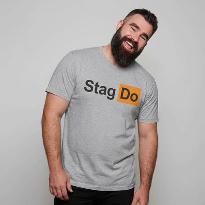 A bearded man in a gray Stag Do PornHub Tee smiles and tilts his head to the side against a plain light background, the humorous shirt adding a playful touch to his relaxed and cheerful vibe.