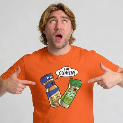 A man points at his "I’m Cumin!" Stag Do Tee featuring cartoon spice jars—Ground Cumin saying "I'm Cumin!" and a surprised Herb Blend—while looking upward with a humorous expression.
