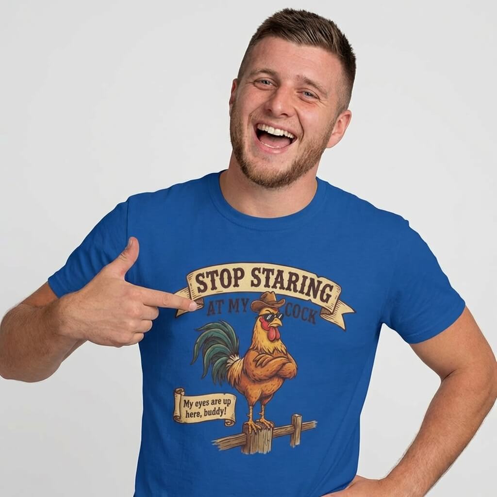 A smiling man points to his blue Stop Staring at My Cock Stag Do Tee, featuring a cartoon rooster and humorous text—ideal for stag parties or as a funny groom t-shirt—standing against a plain light background.