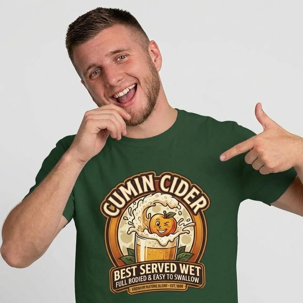A smiling man in a green shirt points at his chest, wearing the Cumin Cider Stag Do Tee. This funny shirt shows a cider glass, cartoon apple, and the slogan: "Cumin Cider: Best served wet. Full bodied & easy to swallow.