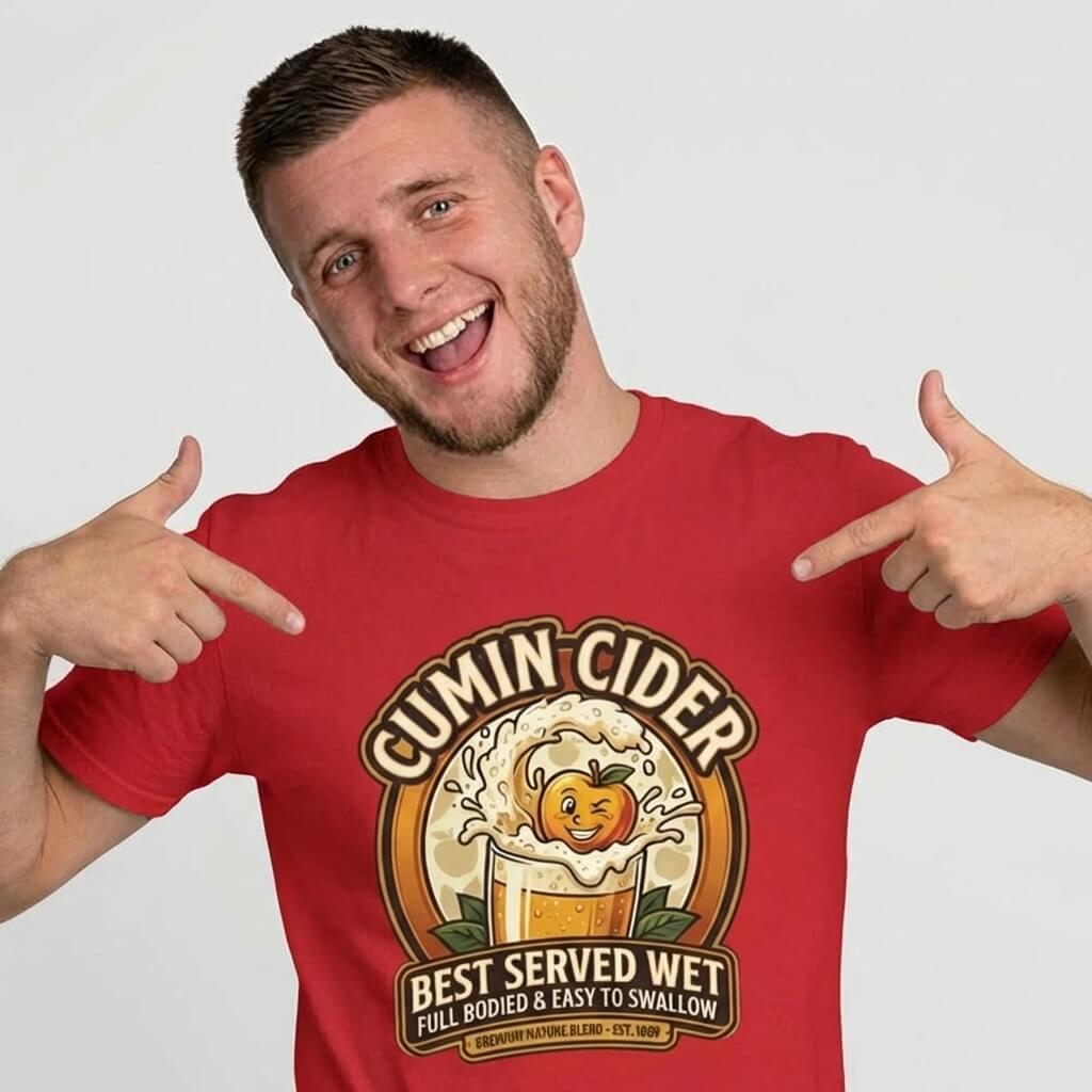 A smiling man with short hair wears the Cumin Cider Stag Do Tee, featuring a frothy cider glass and cartoon apple. He points at the shirt, which says "Best Served Wet, Full Bodied & Easy to Swallow"—an ideal groom gift!.