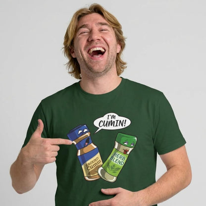 A man with shoulder-length blond hair laughs and points at his green "I’m Cumin!" Stag Do Tee featuring cartoon spice jars, one labeled Ground Cumin saying "I’m Cumin!" and another labeled Herb Blend.