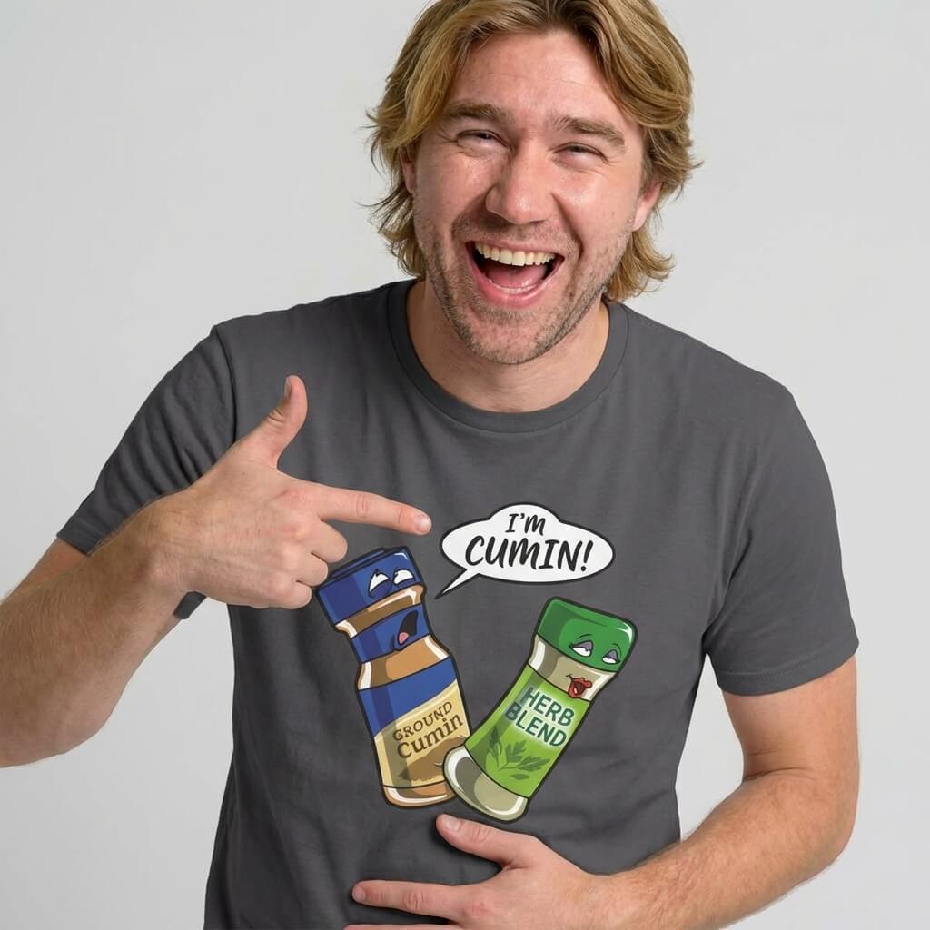 A smiling man with medium-length blond hair points at his "I’m Cumin!" Stag Do Tee, featuring cartoon spice jars—one labeled Ground Cumin saying "IM CUMIN!" to another jar labeled Herb Blend.