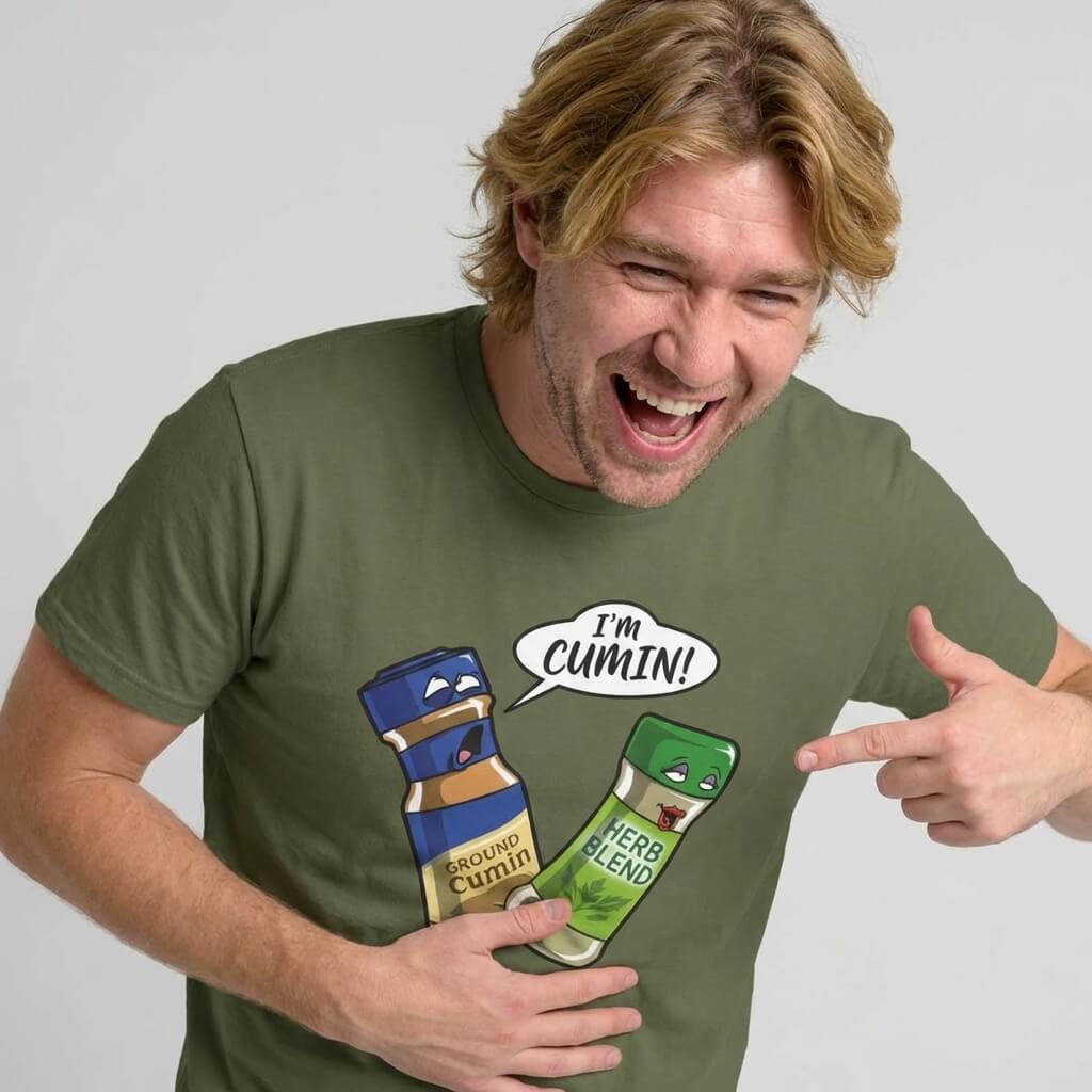 A laughing man wears the "I’m Cumin!" Stag Do Tee—an olive green shirt with cartoon spice bottles. One bottle, Ground Cumin, looks shocked as Herb Blend exclaims “IM CUMIN!”—the perfect funny stag do shirt.