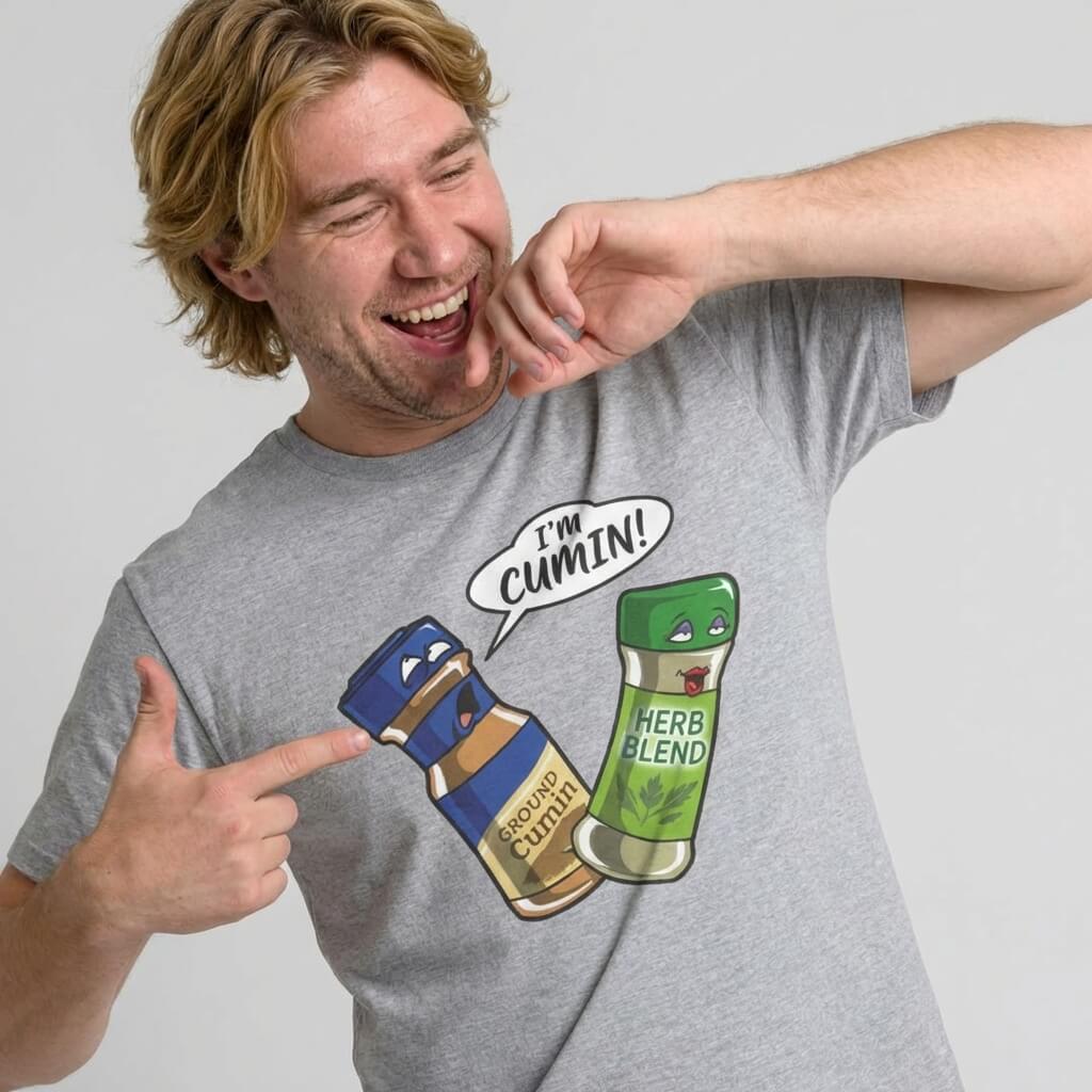 A smiling man points at his gray "I’m Cumin!" Stag Do Tee, featuring cartoon bottles of ground cumin and herb blend. The cumin bottle says, “I’m cumin!”—a hilarious option for a funny stag do shirt.