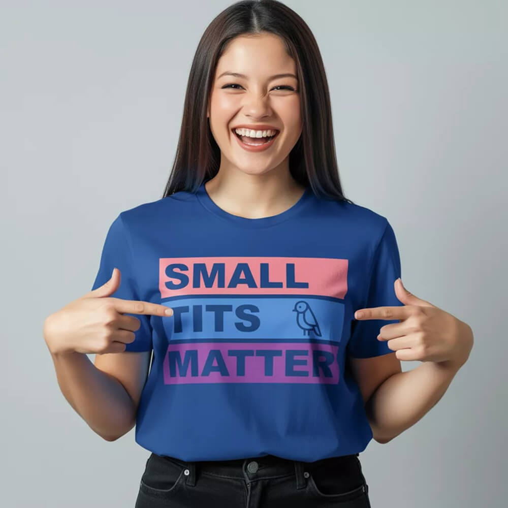 A smiling woman with long dark hair points at her "Small Tits Matter" Tee in blue, featuring bold text and a small bird icon—an empowering women's shirt ideal for making a statement.