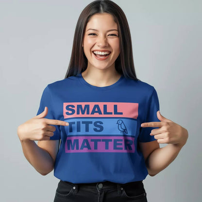 A smiling woman with long dark hair points at her "Small Tits Matter" Tee in blue, featuring bold text and a small bird icon—an empowering women's shirt ideal for making a statement.
