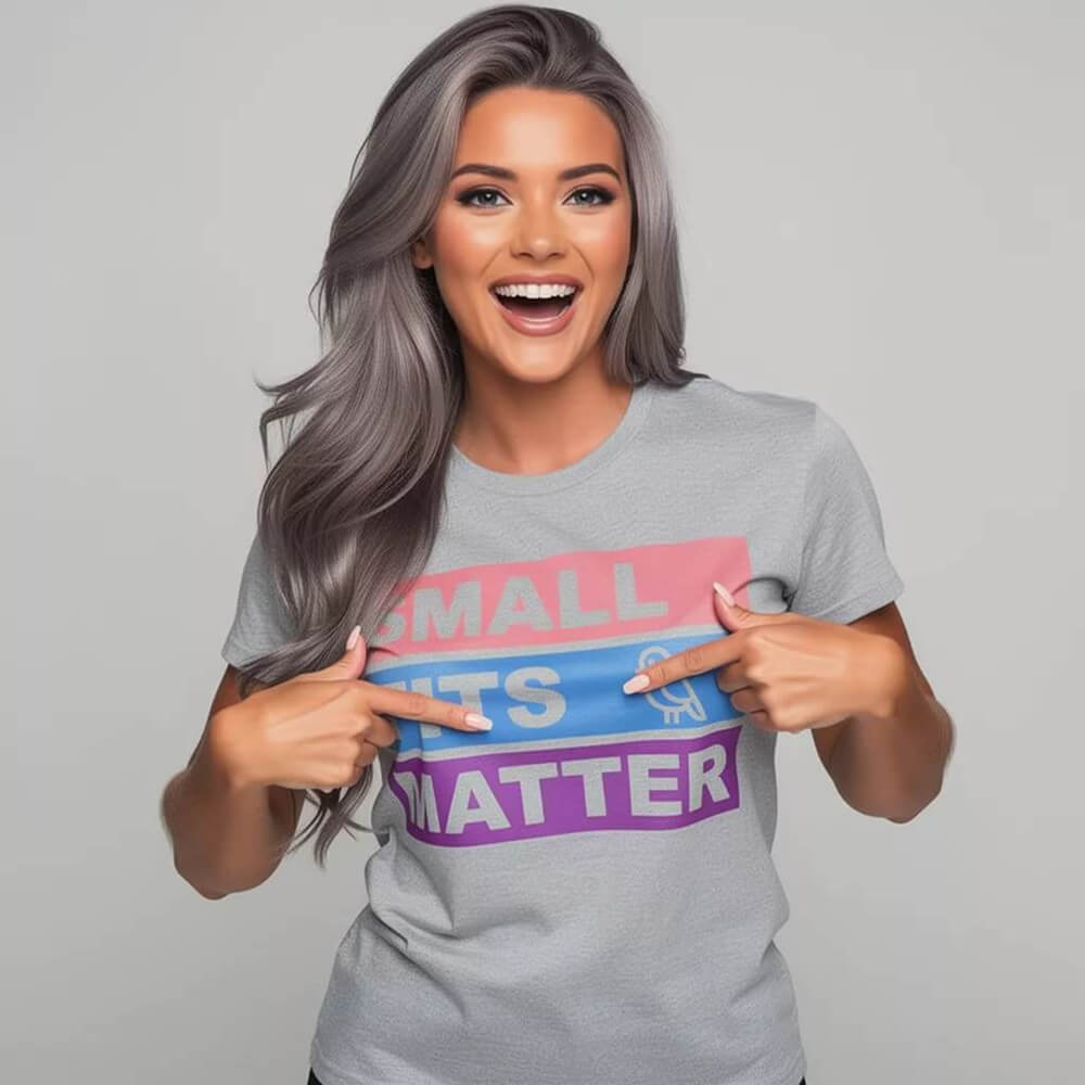 A smiling woman with long gray hair points at her empowering "Small Tits Matter" Tee—a light gray hen do t-shirt with colorful block text in pink, blue, and purple—set against a plain, light background.
