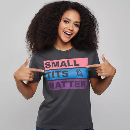 A smiling woman with curly hair points at her "Small Tits Matter" Tee, which features colorful blocks and a small bird graphic. She stands against a plain light background, proudly showcasing this empowering shirt for women.