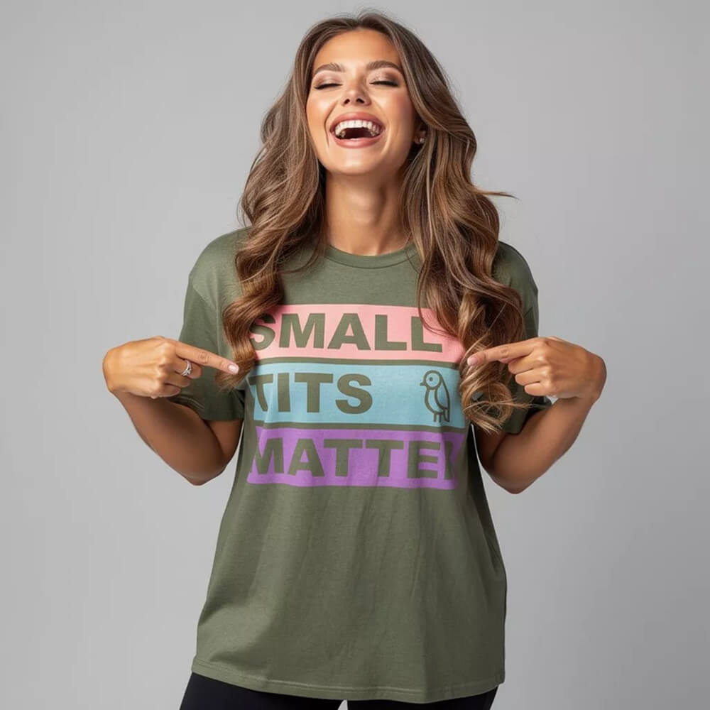 A smiling woman with long brown hair points at her "Small Tits Matter" Tee—an olive green shirt featuring colorful text and a small bird graphic. The background is plain gray.