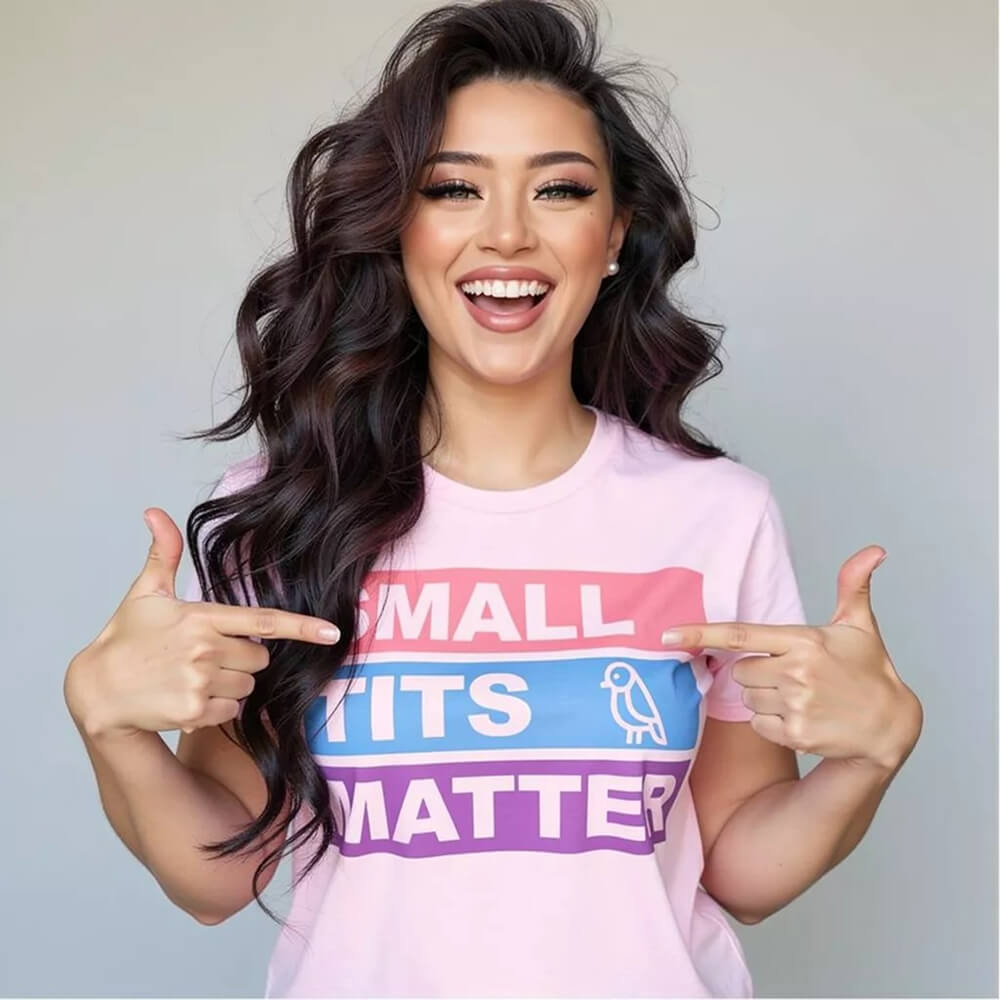 A smiling woman with long wavy hair wears the "Small Tits Matter" Tee, featuring colorful block letters and a bird icon for the I in TITS. She points at the playful text—adding fun to this cheeky statement shirt!.