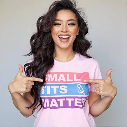 A smiling woman with long wavy hair wears the "Small Tits Matter" Tee, featuring colorful block letters and a bird icon for the I in TITS. She points at the playful text—adding fun to this cheeky statement shirt!.