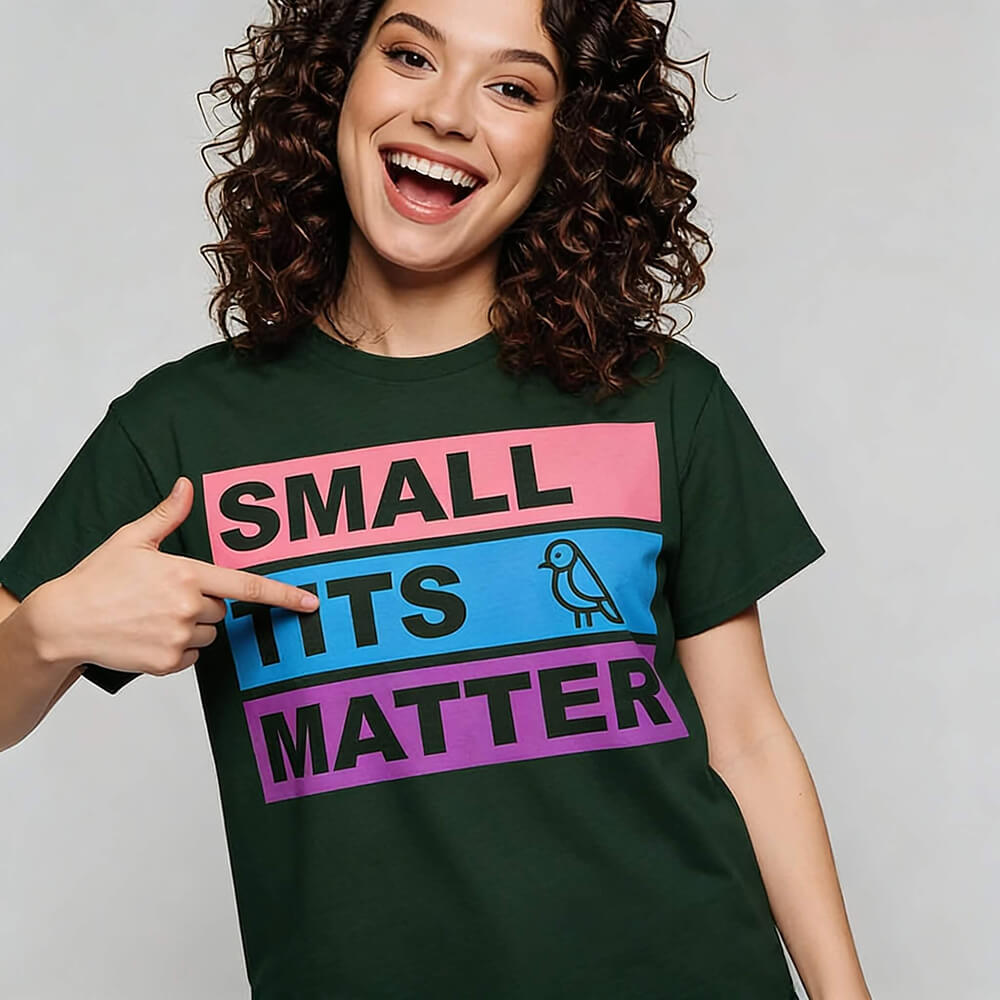 A smiling person with curly hair points to their "Small Tits Matter" Tee—a dark green shirt featuring colorful blocks, the phrase SMALL TITS MATTER, and a small bird illustration on the center block.