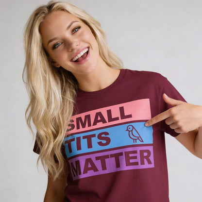 A smiling woman with long blonde hair points to her maroon "Small Tits Matter" Tee, which features bold text and a small bird graphic.