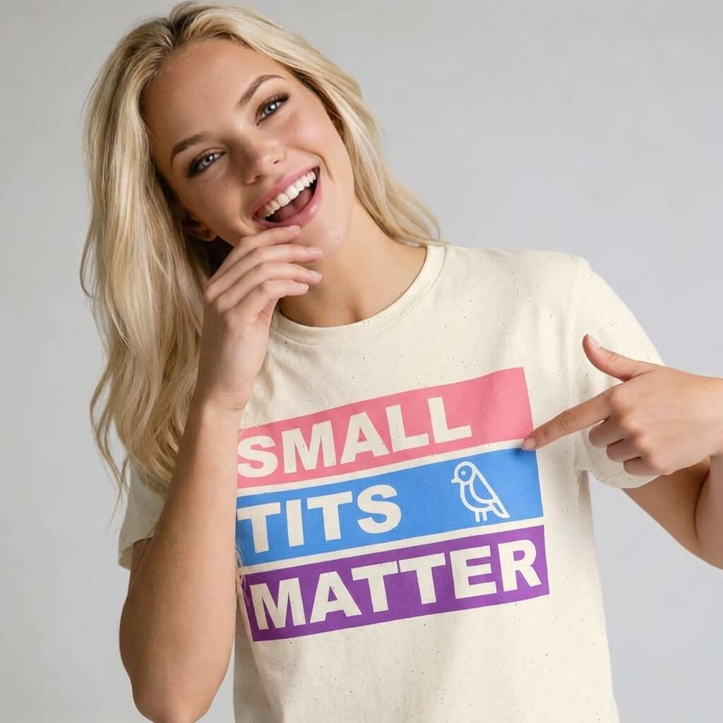 A smiling woman with long blonde hair points to her "Small Tits Matter" Tee, featuring colorful blocks and a playful bird—an empowering shirt for women who want to make a bold statement.