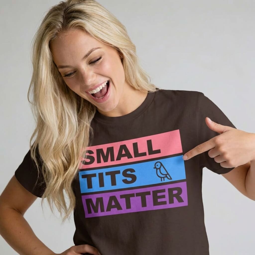 A smiling woman with long blonde hair points to her "Small Tits Matter" Tee, featuring bold lettering and a small bird icon, set against a colorful striped background.