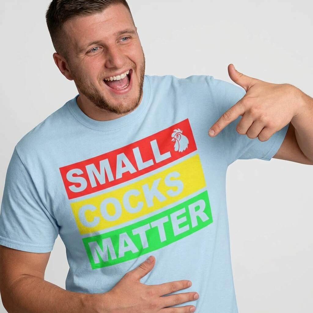 A smiling man wears the Small Cocks Matter Stag Do Tee, pointing at its bold red, yellow, and green text while holding his stomach—an ideal choice for those seeking humorous stag do shirts.