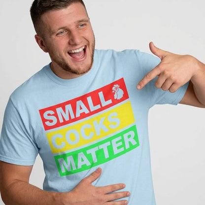 A smiling man wears the Small Cocks Matter Stag Do Tee, pointing at its bold red, yellow, and green text while holding his stomach—an ideal choice for those seeking humorous stag do shirts.