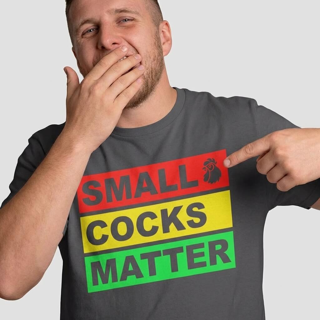 A man smiles, hand over mouth, and points to his Small Cocks Matter Stag Do Tee—a body confidence shirt with bold red, yellow, and green text plus a small cartoon rooster beside the word SMALL.