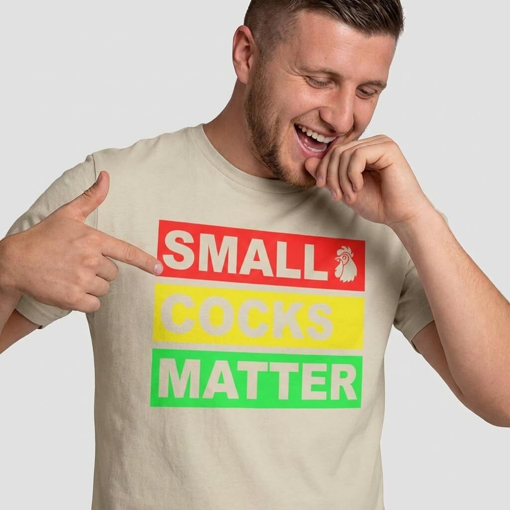 A man smiles and points to his beige Small Cocks Matter Stag Do Tee, a funny bachelor party shirt with bold red, yellow, and green text plus a rooster graphic by the word SMALL.