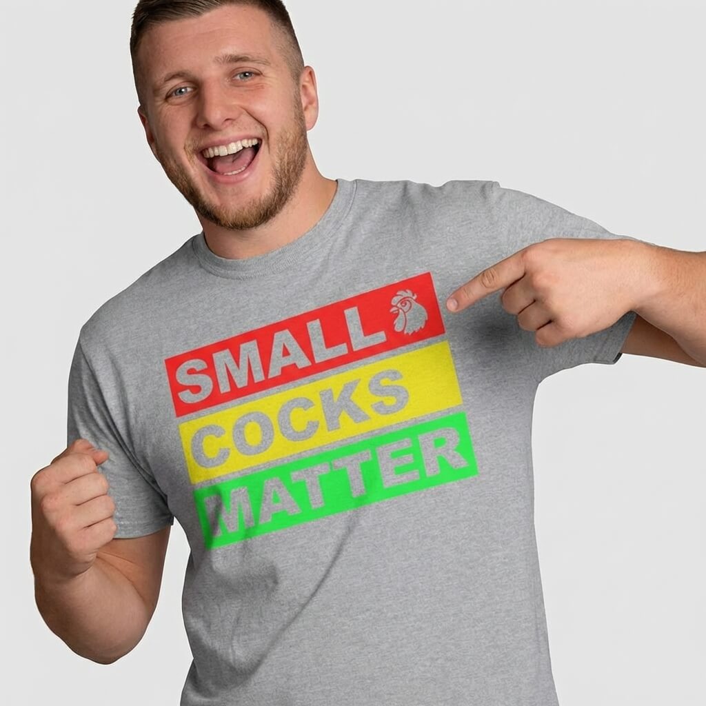 A smiling man points to his gray Small Cocks Matter Stag Do Tee, featuring bold red, yellow, and green text blocks with a rooster icon—perfect for bachelor party laughs and making a statement.