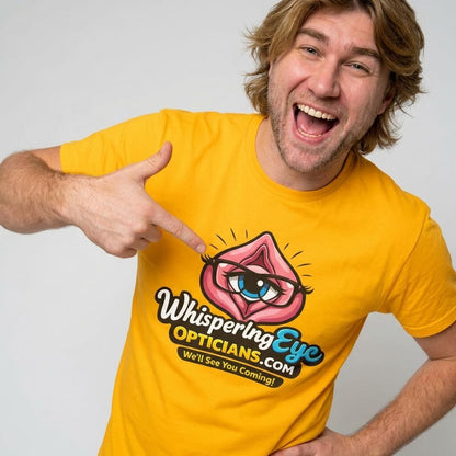 A smiling man with shoulder-length hair wears the bright yellow "Whispering Eye Opticians Logo Tee (We'll See You Coming!)", pointing enthusiastically at the fun stag do shirt featuring an eye inside pink lips.