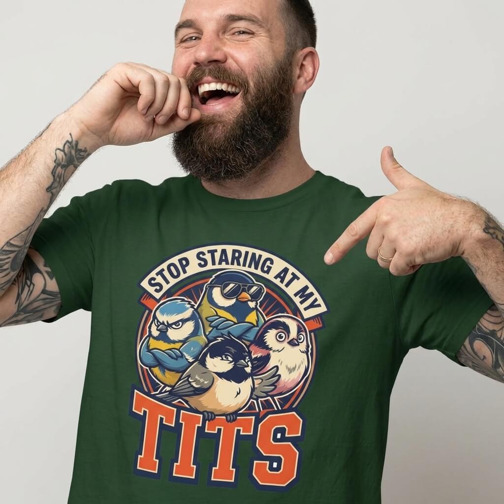 A bearded man with tattooed arms smiles and points at his “Stop Staring at My Tits” Stag Do Tee, featuring cartoon birds—a hilarious choice for funny stag do outfits.