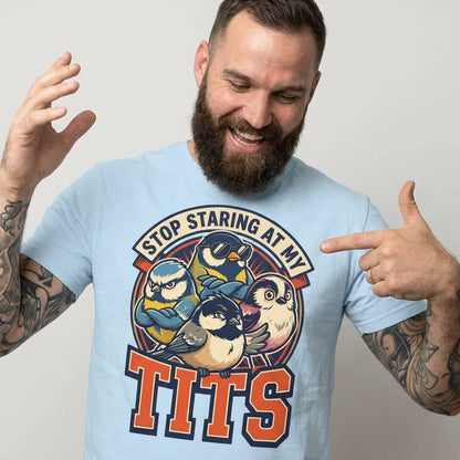 A bearded man with tattoos points at his light blue “Stop Staring at My Tits” Stag Do Tee, featuring cartoon birds and bold text—an ideal choice for hilarious stag do outfits or a unique gift for the groom.