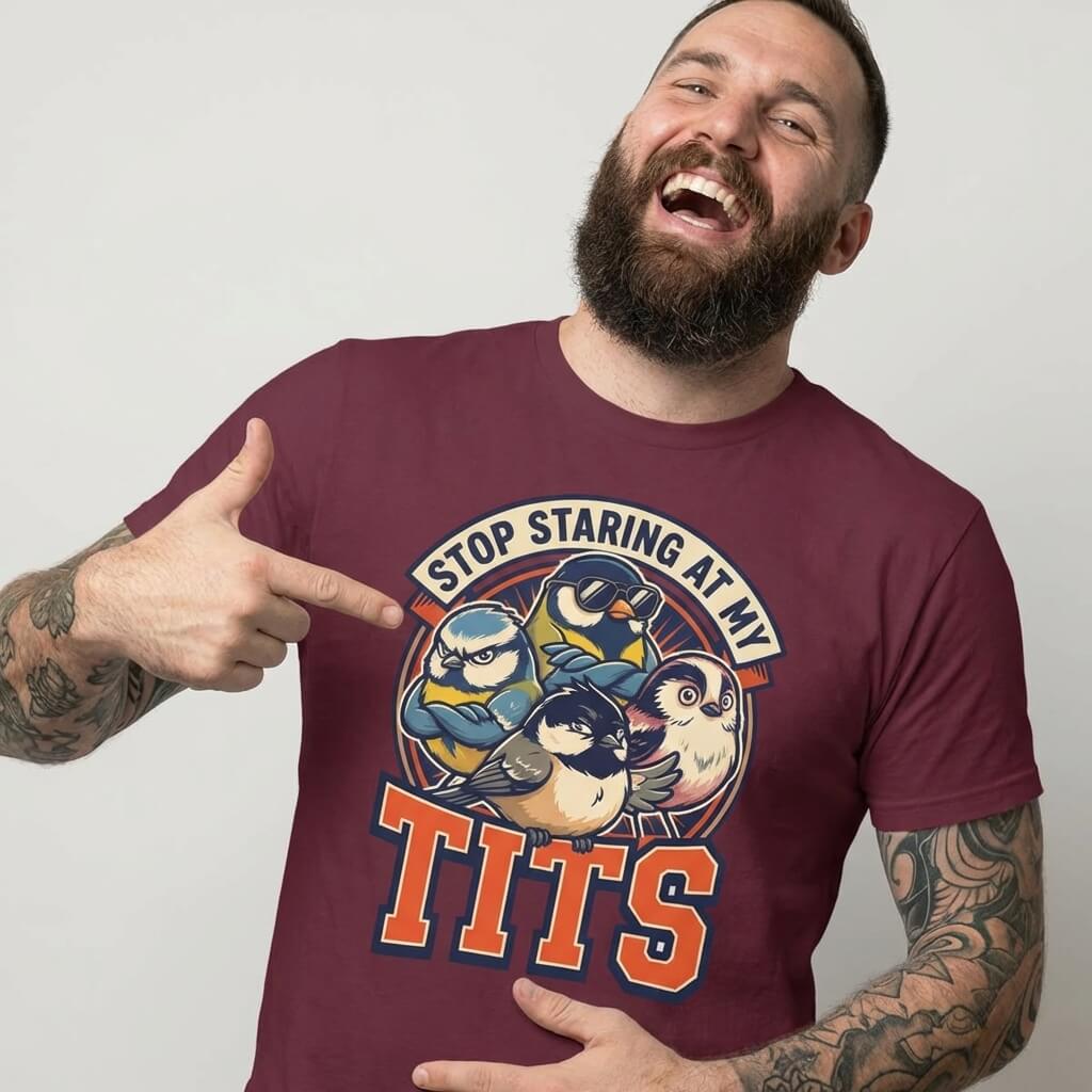A bearded, tattooed man smiles and points to his “Stop Staring at My Tits” Stag Do Tee with cartoon birds—a hilarious choice for stag do outfits or a unique gift for the groom.