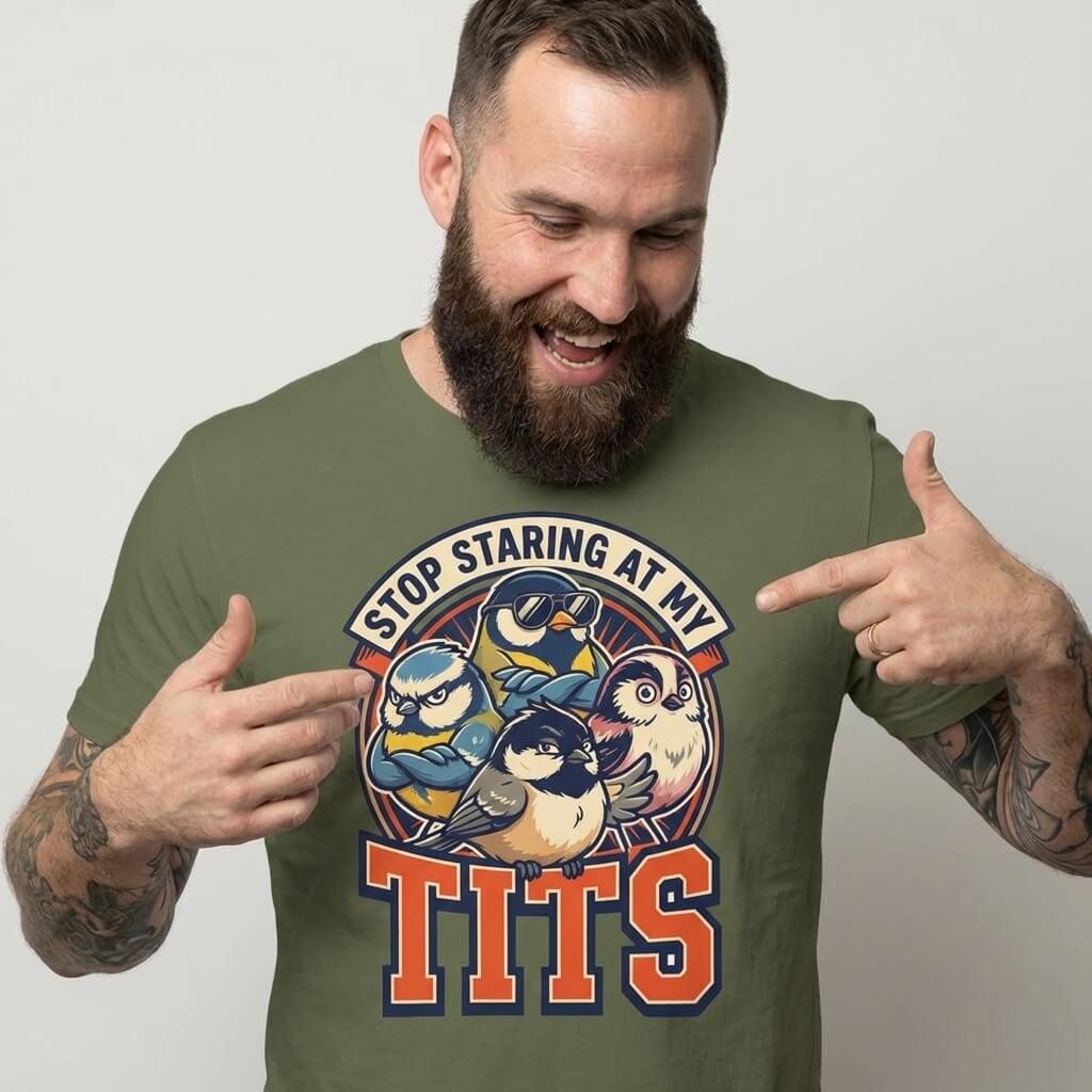 A tattooed, bearded man smiles and points at his green “Stop Staring at My Tits” Stag Do Tee, a hilarious shirt with cartoon birds—perfect for funny stag parties or as a groom gift.