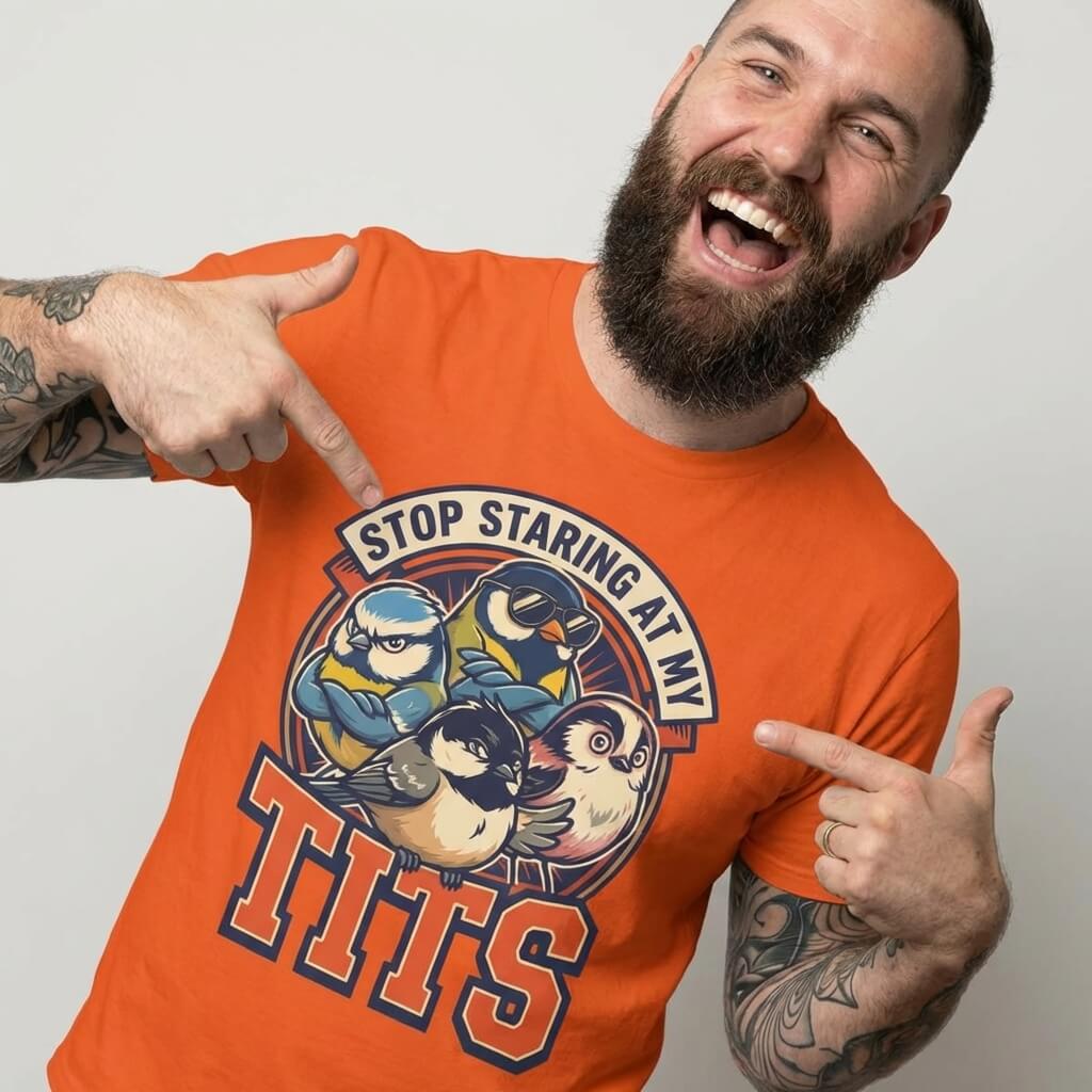 A bearded man in an orange “Stop Staring at My Tits” Stag Do Tee with cartoon birds smiles and points to his shirt—a funny gift for the groom or stag do.