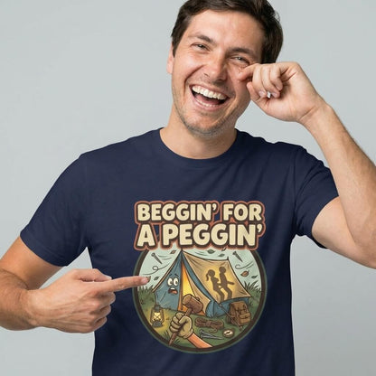 A smiling man points to his Beggin' for a Peggin' Camping Tee, featuring a cartoon campsite and silhouetted tent figures. This funny, rude t-shirt is perfect as a cheeky gift or for adding humor to any festival outfit.