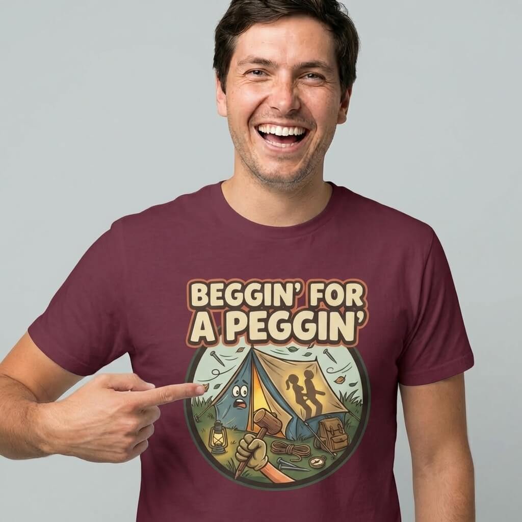 A smiling man in a maroon Beggin' for a Peggin' Camping Tee points to the shirt, which shows a cartoon campsite scene and funny text—perfect for festival outfits or as a cheeky gift guaranteed to get laughs.