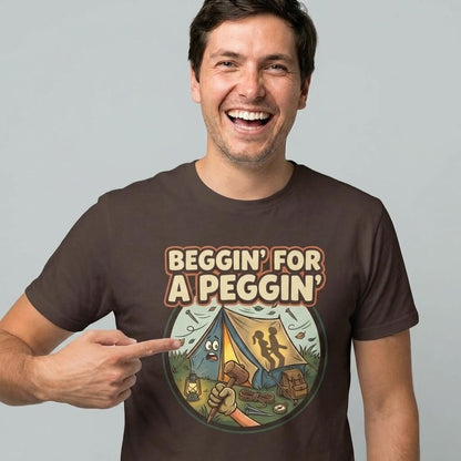 A smiling man points to his Beggin' for a Peggin' Camping Tee featuring a cartoon campsite and the bold text “Beggin’ for a Peggin’”—the perfect rude gift or standout festival outfit.