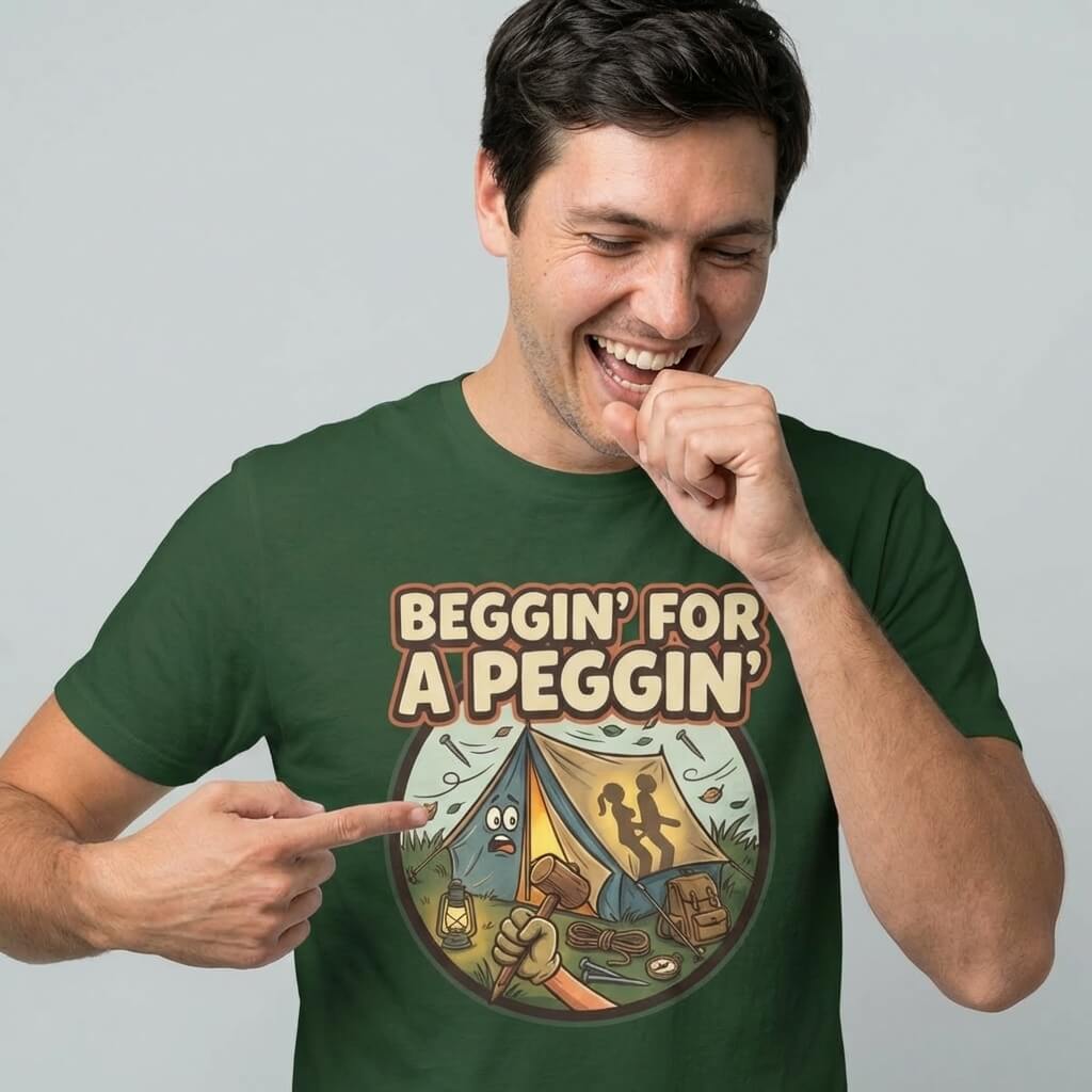 A man laughs and points at his green Beggin' for a Peggin' Camping Tee, featuring a colorful cartoon tent scene—an ideal cheeky festival outfit or funny, rude gift.