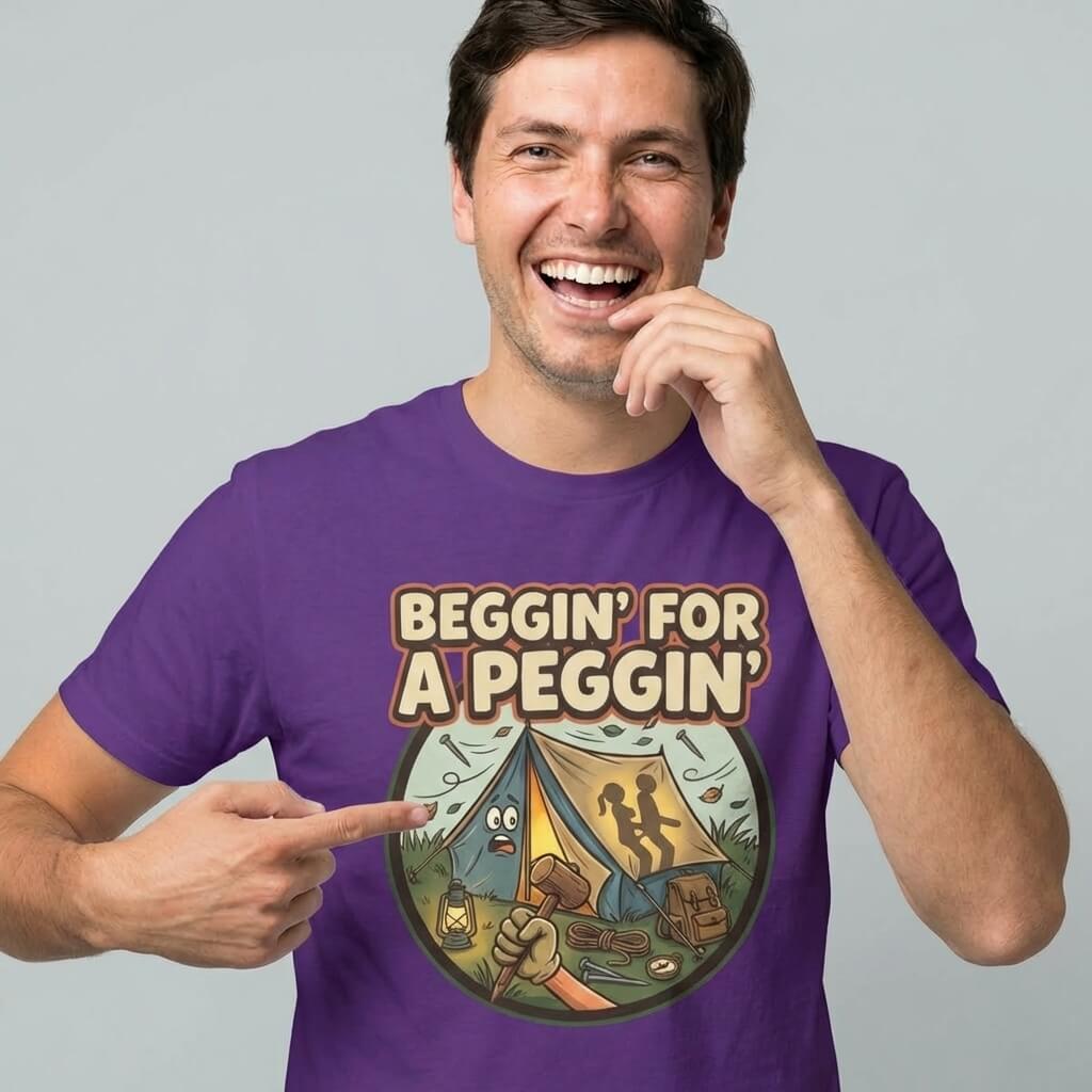 A smiling man wears the Beggin' for a Peggin' Camping Tee, a purple shirt with a cartoon camp scene and two silhouettes in a tent—an ideal rude gift or quirky festival outfit.