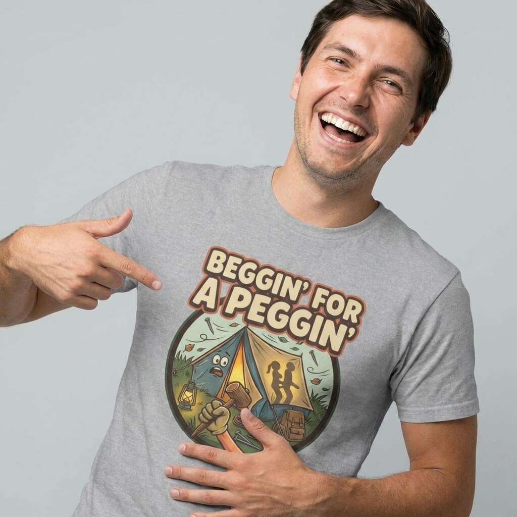A smiling man in a gray Beggin' for a Peggin' Camping Tee points to his shirt, which features a cartoon tent, camping gear, and the text BEGGIN FOR A PEGGIN—a hilarious gift or funny camping T-shirt for festivals.
