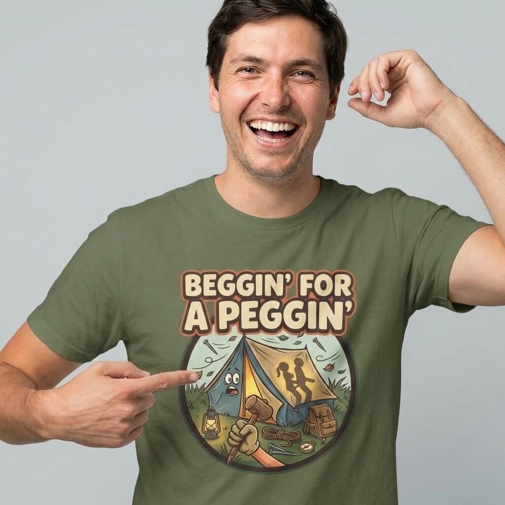 A smiling man points to his green Beggin' for a Peggin' Camping Tee, featuring two silhouetted figures in a tent and the text "Beggin for a Peggin"—the perfect rude festival or camping gift.