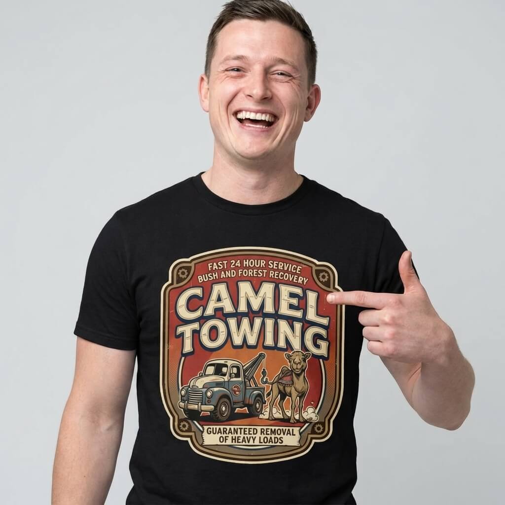 A smiling man points to his black Camel Towing Funny Tee, featuring a colorful camel and tow truck graphic with the text: "Fast 24 hour service, guaranteed removal of heavy loads"—a perfect novelty gift.